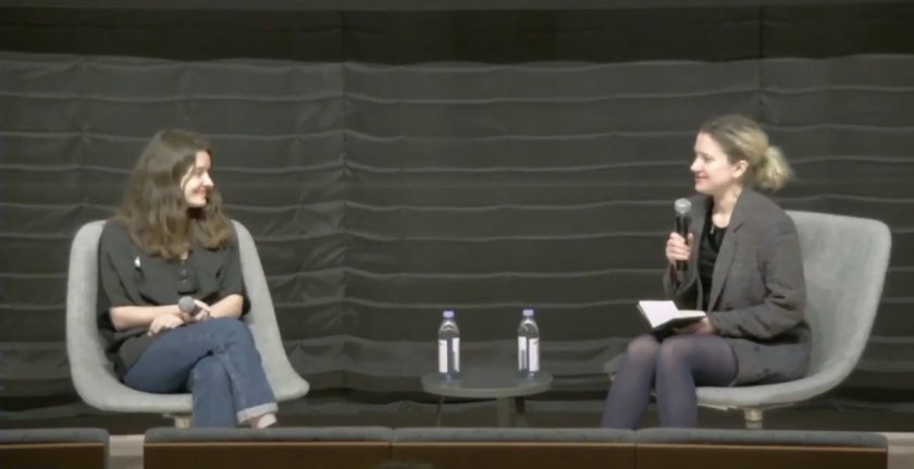 A screenshot from a video of the event, Anna Tropnikova and Ukrainian filmmaker Iryna Tsilyk sit in chair's facing one another on a dark grey stage. The women are smiling and holding microphones.